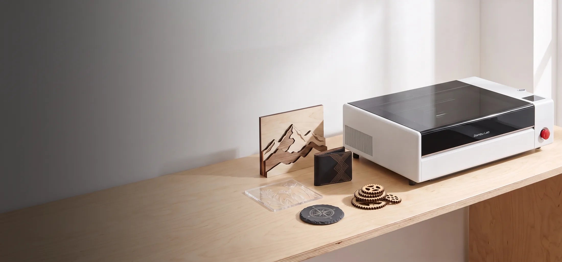 Bambu Lab R1 CO2 laser cutter on a birch workbench with laser-cut wood art, an engraved leather wallet, an acrylic panel and a slate coaster