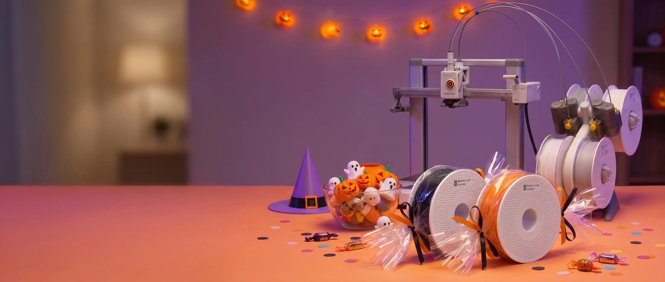 Bambu Lab A1 Combo on a party table beside a bowl of small 3D-printed pumpkins and ghosts, with two Bambu Lab PLA Basic spools in black and pumpkin orange